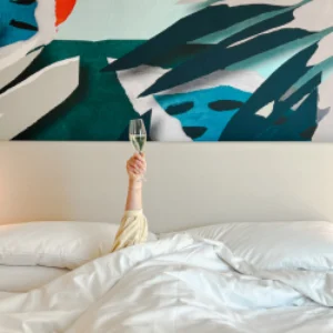 Guest celebrating with a glass of champagne in bed, surrounded by crisp white linens and a bold artistic mural — luxury hotel marketing and hospitality branding inspiration